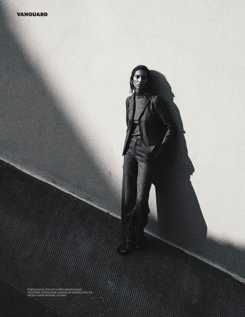 Model wearing a dark tailored suit leans against a textured wall under hard directional lighting in black and white.