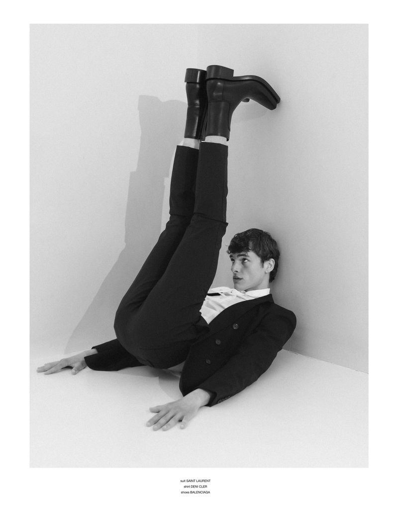 Model Dominik Schmidt lying on the floor in a black suit and boots with legs extended upward against a corner wall.