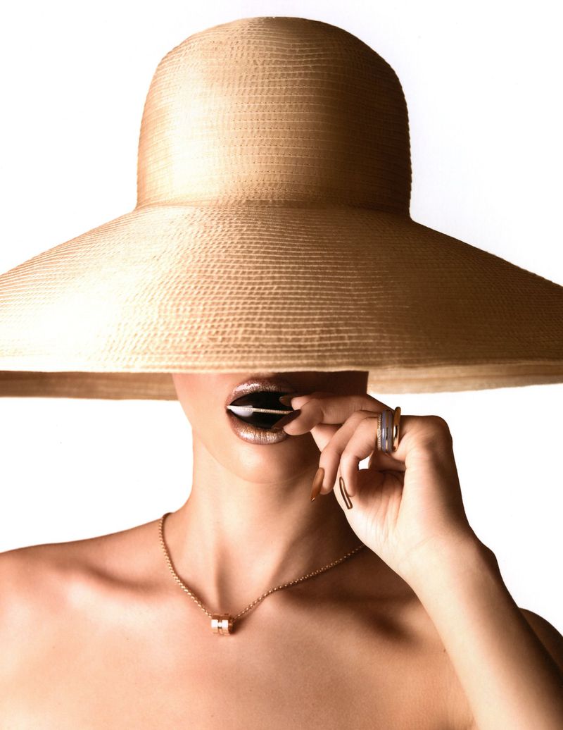 Woman wearing an oversized beige woven sunhat over her eyes, with metallic lips, gold jewelry, and bare shoulders on a white background.