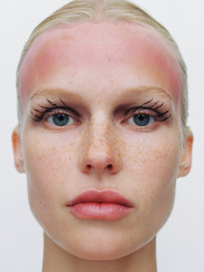 Close-up portrait of a woman with freckles, graphic drawn eyelashes, and a pink gradient across her forehead on a white background.