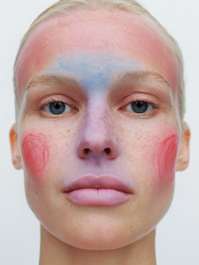Close-up beauty portrait of a blonde woman with blue eyes featuring pastel red, pink, and purple face paint on a white background.