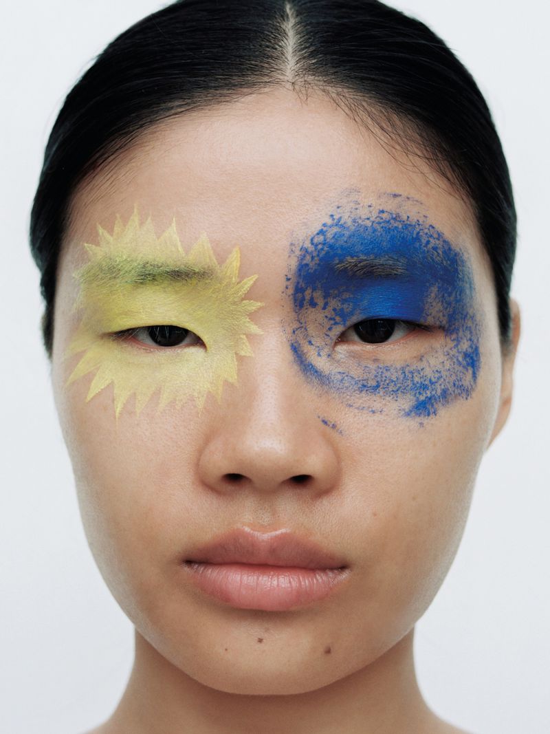 Close-up portrait of an East Asian woman with graphic yellow and smudged blue eye makeup against a white studio background.