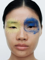 Close-up portrait of an East Asian woman with graphic yellow and smudged blue eye makeup against a white studio background.