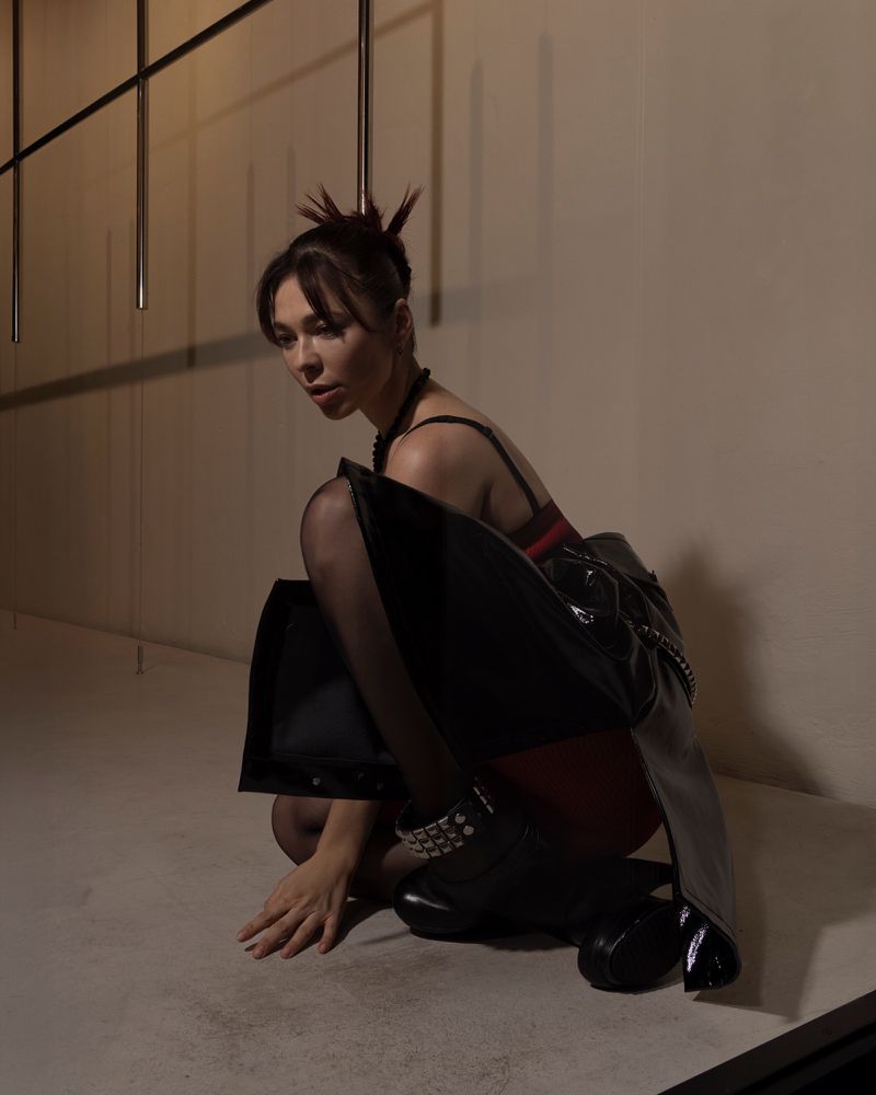Nina Kraviz crouching in a structured black leather garment, sheer tights, and studded boots against a studio wall.