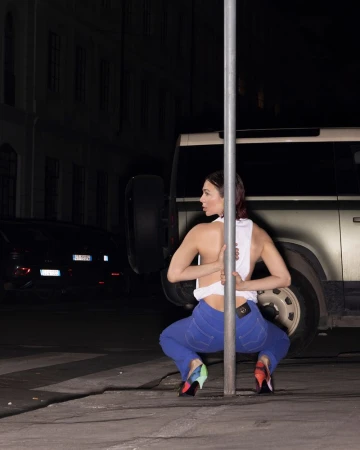 Nina Kraviz crouches behind a metal pole in blue jeans, a white tank top, and patterned heels on a dark city street at night.