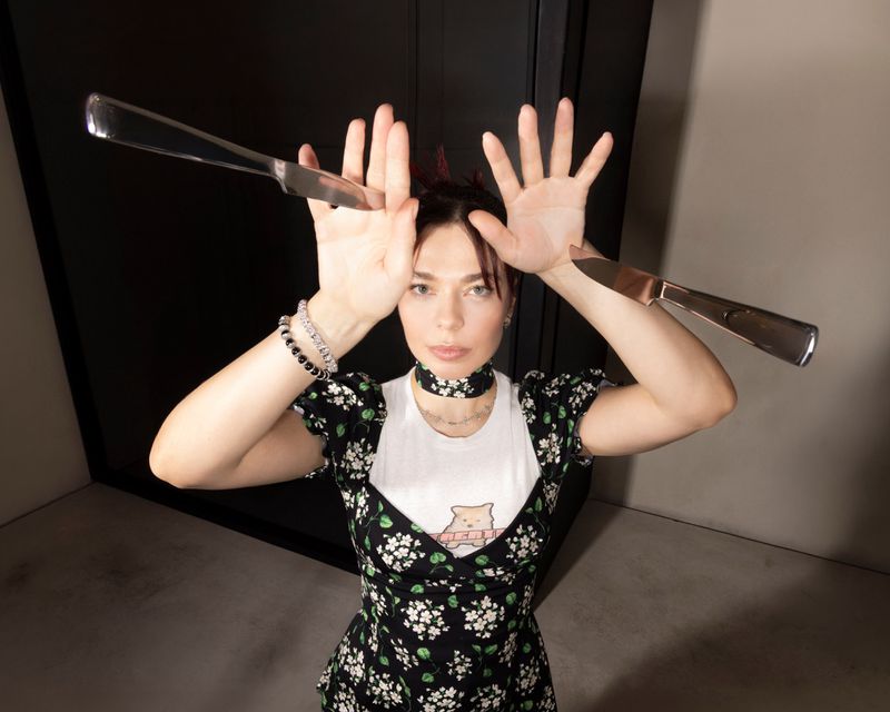 Nina Kraviz holding up open hands balancing silver butter knives wearing a floral print dress over a white top.