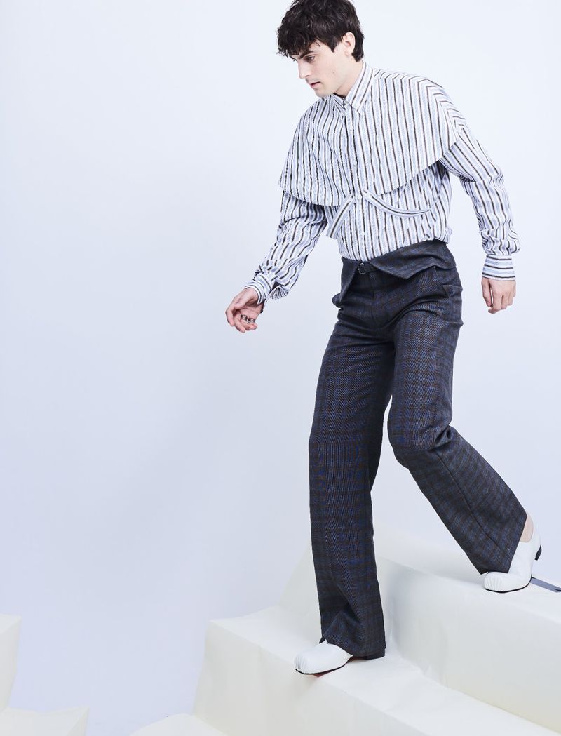Julien De Saint-Jean stepping down white pedestals wearing a striped capelet shirt and dark tailored trousers against a white backdrop.