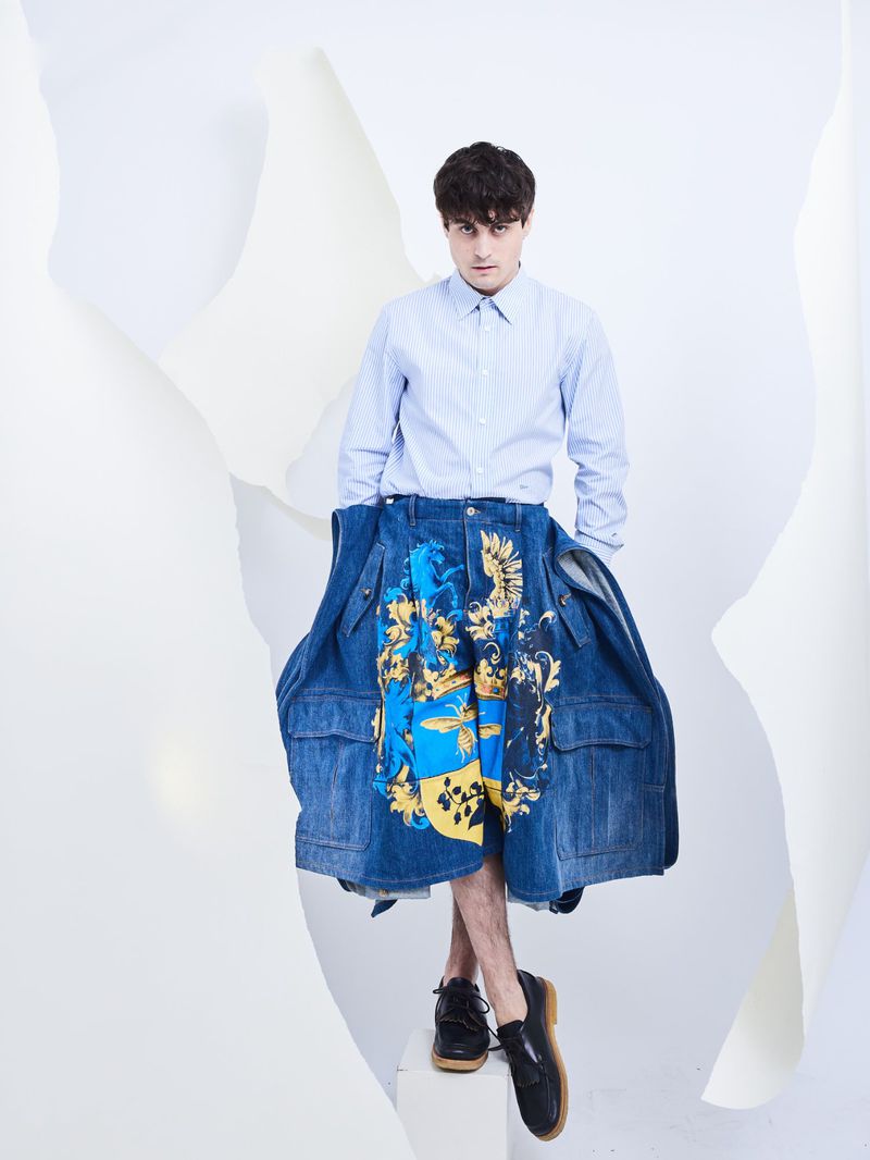 Young man wearing a light blue button-down shirt and oversized printed denim shorts against a white backdrop.