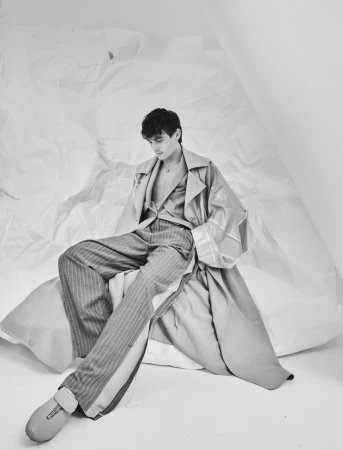 Julien De Saint-Jean seated wearing an oversized coat and pinstripe trousers against a crumpled paper backdrop.