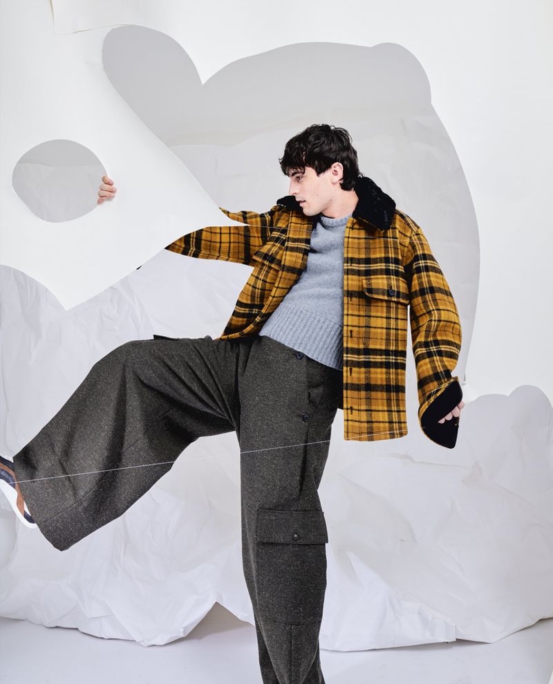 Julien De Saint-Jean in a yellow plaid jacket and wide-leg trousers posing dynamically behind white paper cutouts.