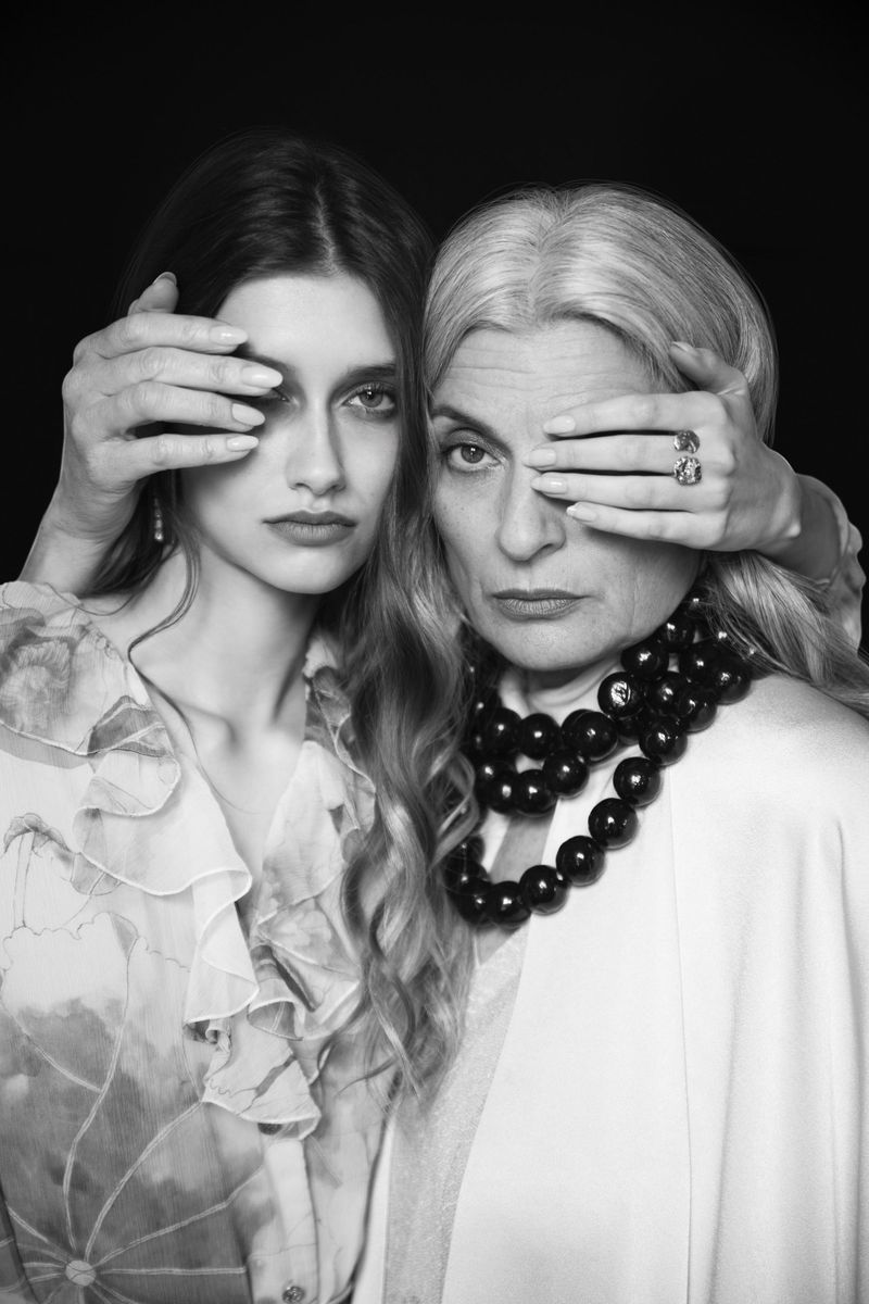 Black and white editorial portrait of two women covering each other's eye with their hands against a dark background.