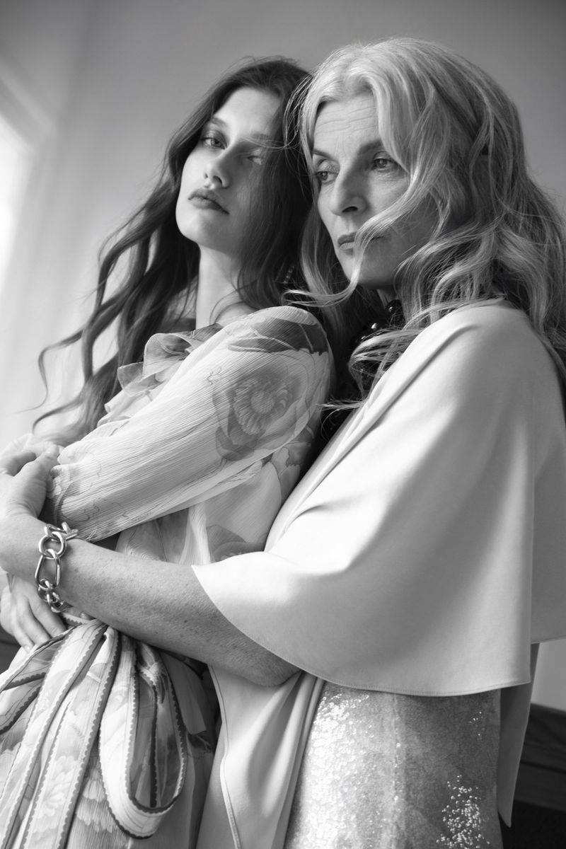 Black and white portrait of two women embracing closely, one in a sheer printed dress and the other in a cape and sequin gown.