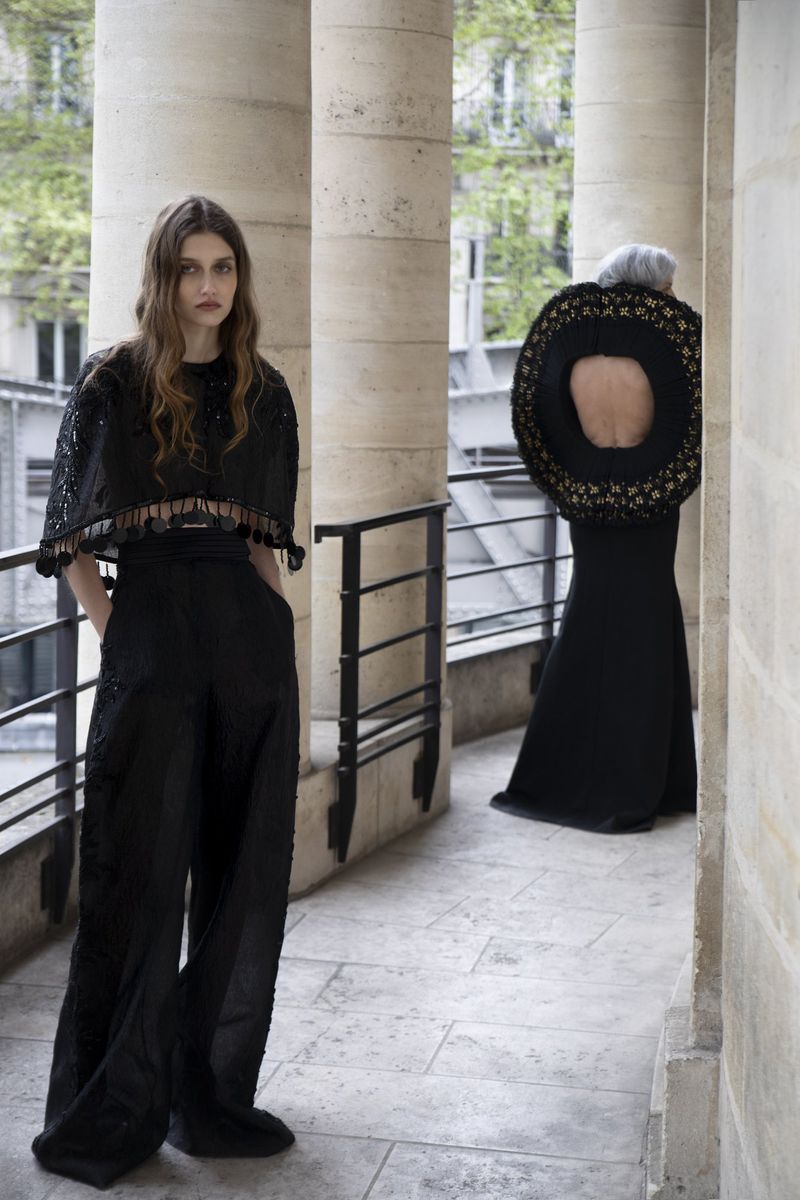 Model wearing a black embellished top and trousers posing on a stone balcony with classical columns.