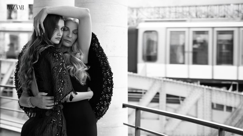 Two women in dark couture embrace beside a stone pillar with a blurred elevated train in the background.