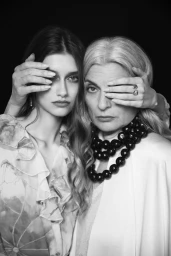 Black and white editorial portrait of two women covering each other's eye with their hands against a dark background.