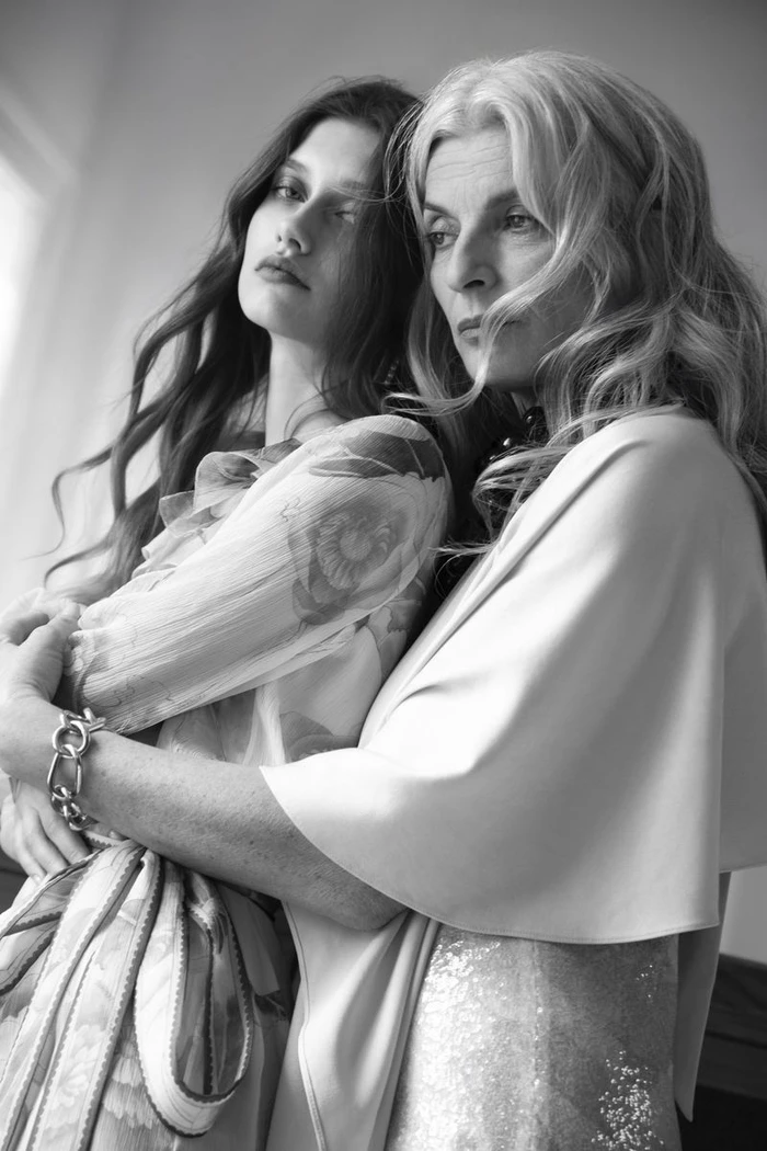 Black and white portrait of two women embracing closely, one in a sheer printed dress and the other in a cape and sequin gown.