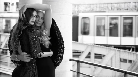 Two women in dark couture embrace beside a stone pillar with a blurred elevated train in the background.