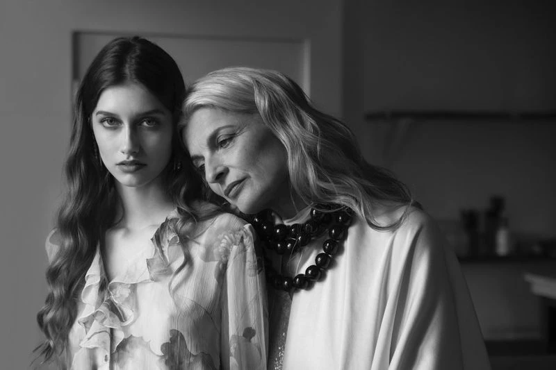 Black and white portrait of a young woman in a ruffled blouse beside a mature woman with a beaded necklace resting on her shoulder.