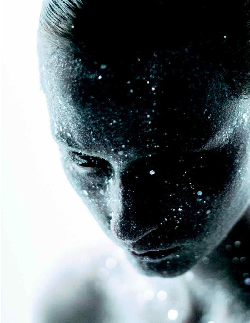 Close-up portrait of model Melinda Kiss looking down with shimmering glitter covering her face against a bright white background.