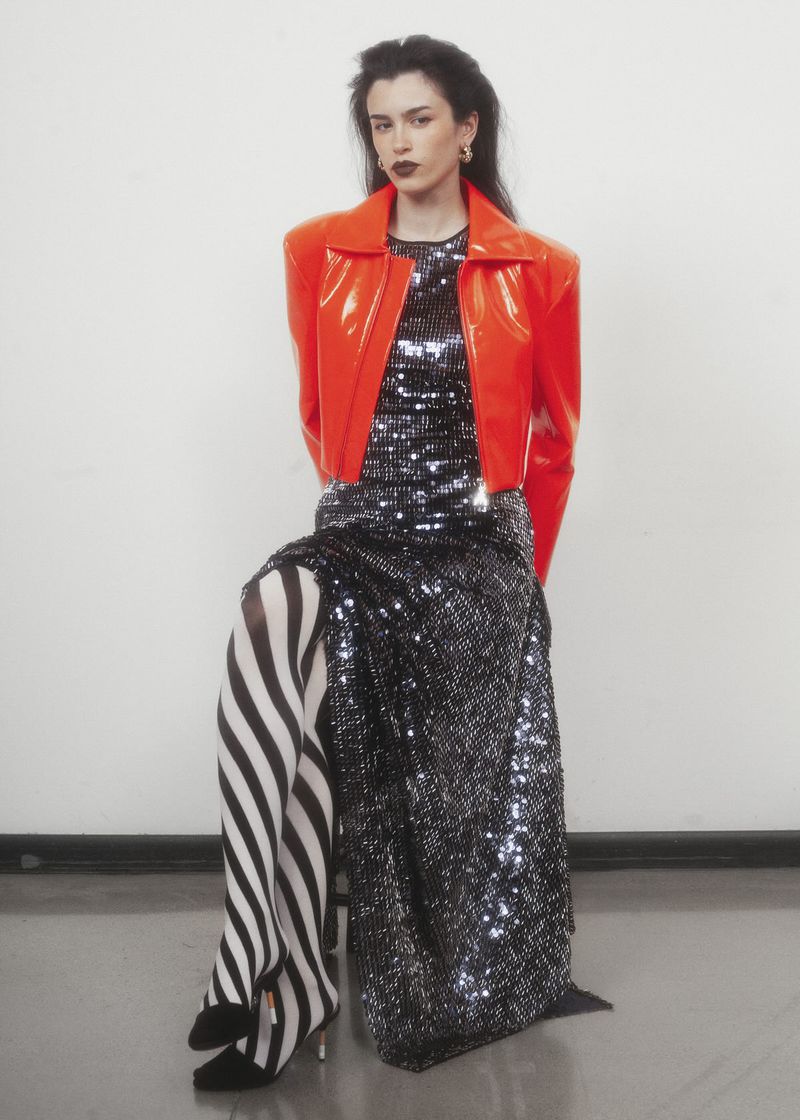 Model wearing a bright orange jacket and sequined gown with black and white striped tights against a plain studio background