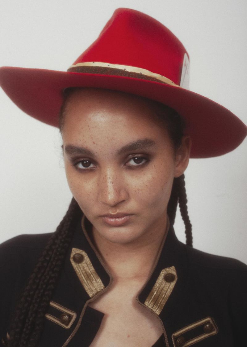 Portrait of a model wearing a wide-brim red hat and a black military-style jacket with gold trim in front of a white backdrop.