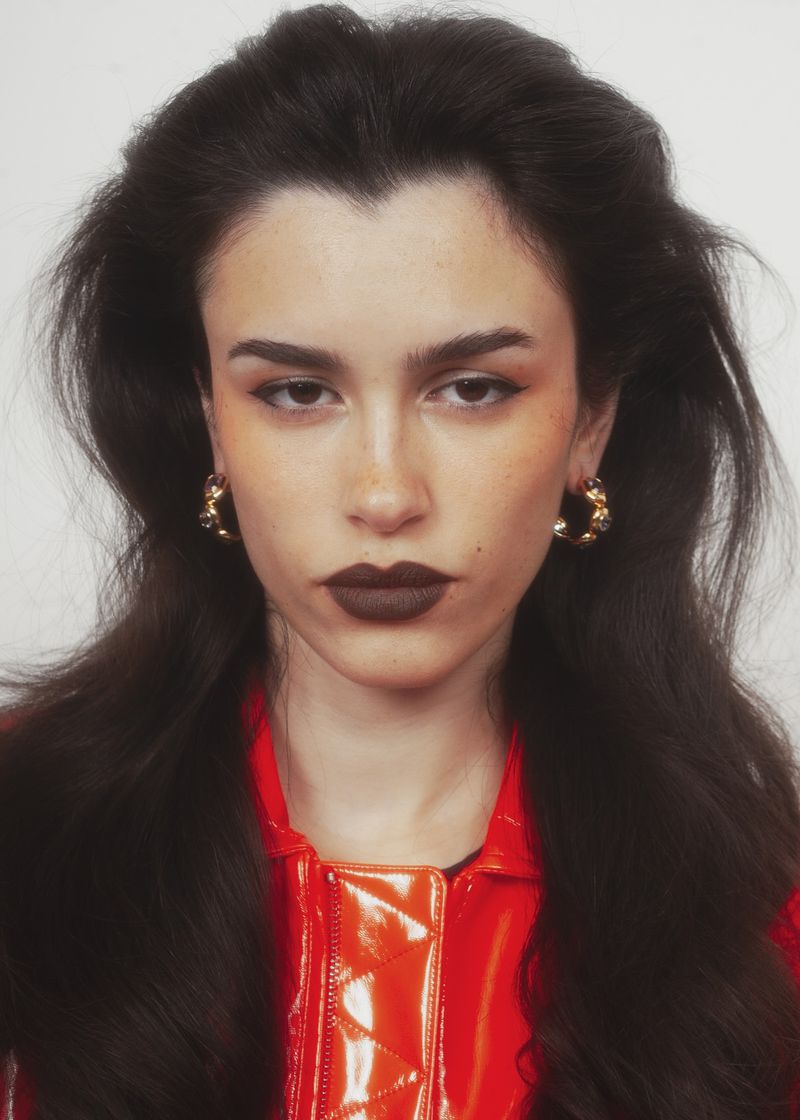 Close-up portrait of a woman with voluminous dark hair, dark lipstick, gold hoop earrings, and a glossy red patent jacket.
