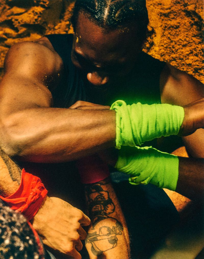Close-up of an athletic Black man with braided hair and neon green wrist wraps looking downward on sand.