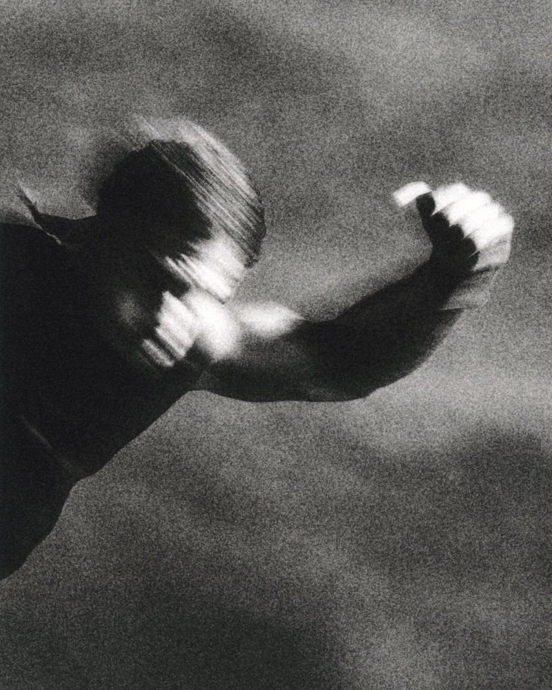 Grainy black and white shot of a shirtless man with motion blur throwing a dynamic punch forward.