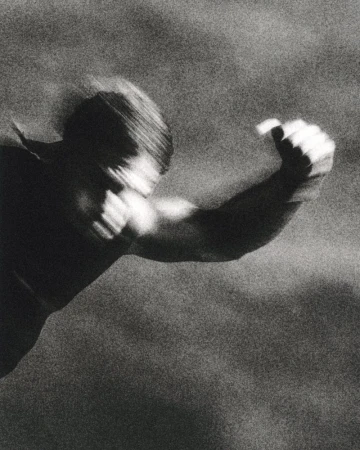 Grainy black and white shot of a shirtless man with motion blur throwing a dynamic punch forward.
