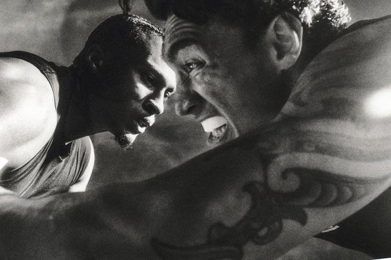 Monochrome close-up of two men confronting each other intensely, one wearing a mouthguard with a prominent arm tattoo.