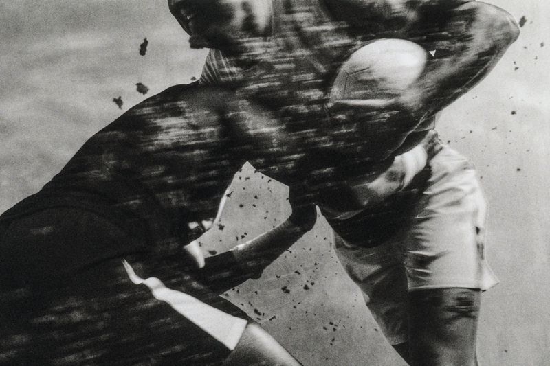 Monochrome photograph of two men in an intense physical tackle during a ball game with motion blur and airborne particles.