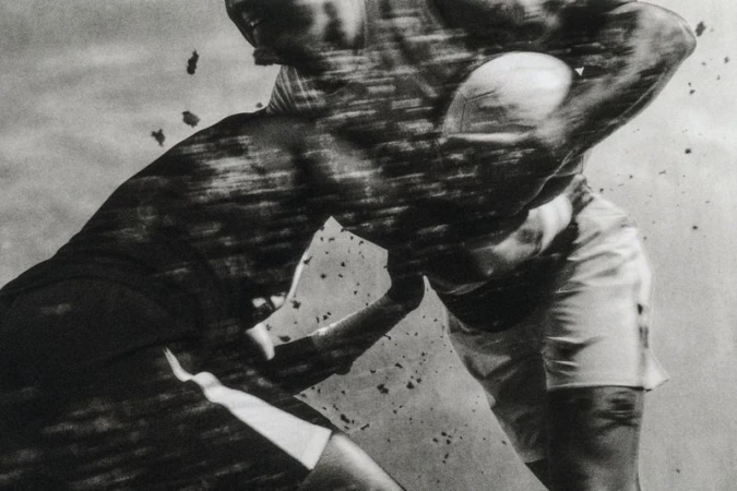 Monochrome photograph of two men in an intense physical tackle during a ball game with motion blur and airborne particles.