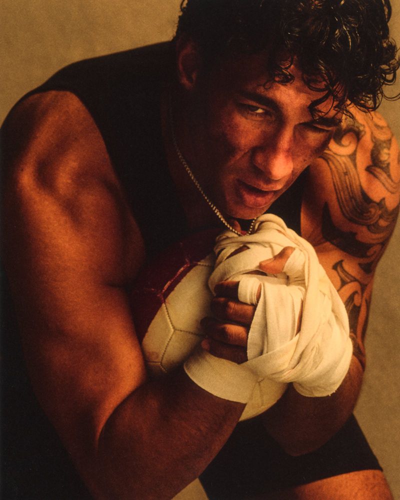 Muscular man with tattooed arm and taped hands tightly clutching a stitched ball against a warm studio backdrop.