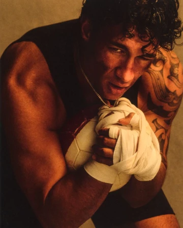 Muscular man with tattooed arm and taped hands tightly clutching a stitched ball against a warm studio backdrop.