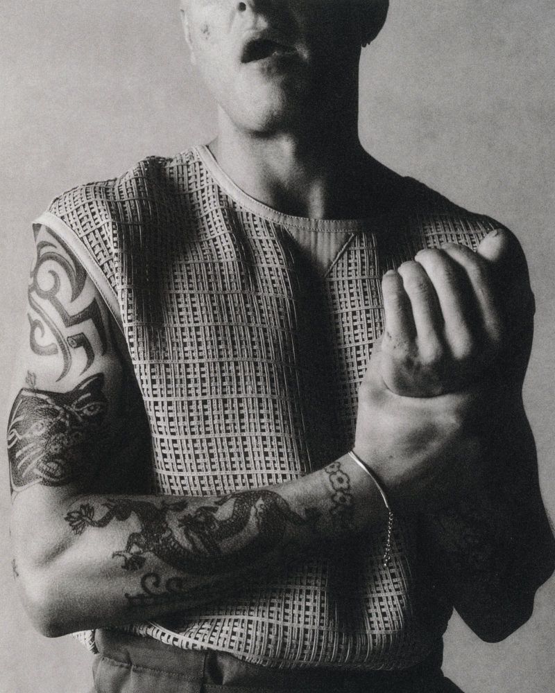 Monochrome portrait of a tattooed man wearing a sleeveless mesh knit top while gripping his wrist against a neutral studio backdrop.