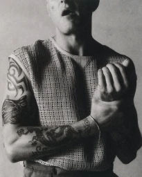 Monochrome portrait of a tattooed man wearing a sleeveless mesh knit top while gripping his wrist against a neutral studio backdrop.
