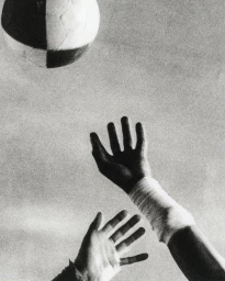 Black and white close-up photograph of hands reaching upward toward a checkered ball in midair with a taped wrist.