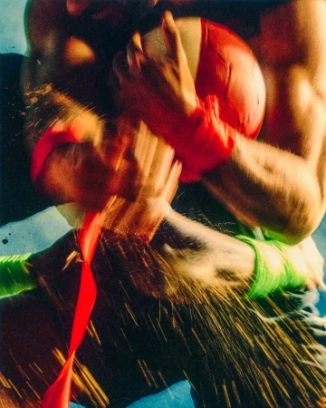 Close-up action shot of two men grappling over a ball with red and neon green wrist wraps amidst motion blur and flying sand.