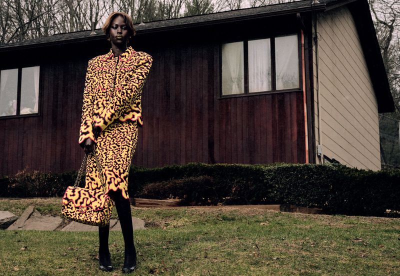Model Aweng Chuol wearing a yellow patterned skirt suit and holding a matching handbag in front of a dark wood house.