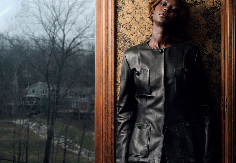 Aweng Chuol posing in a black buttoned leather jacket beside a wood-framed window with patterned wallpaper.