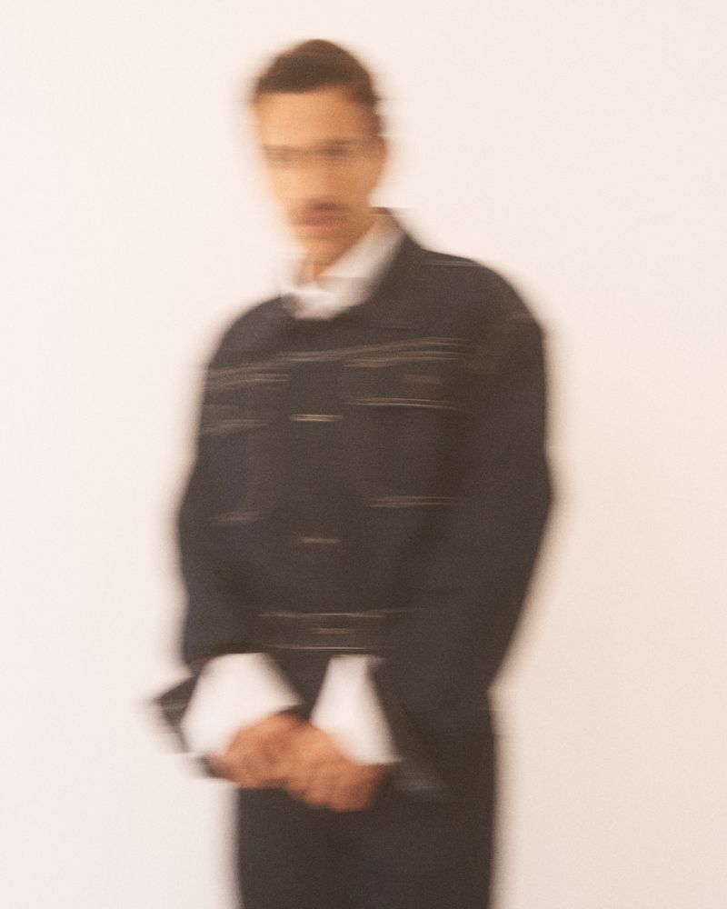 Motion-blurred portrait of a man wearing a dark denim jacket over a white collared shirt against a white seamless backdrop.