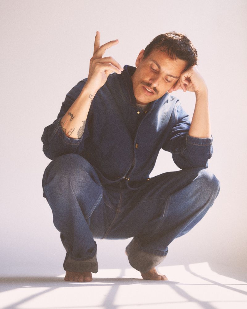 Man with mustache and arm tattoos crouching barefoot wearing a denim jacket and matching jeans in a bright studio.