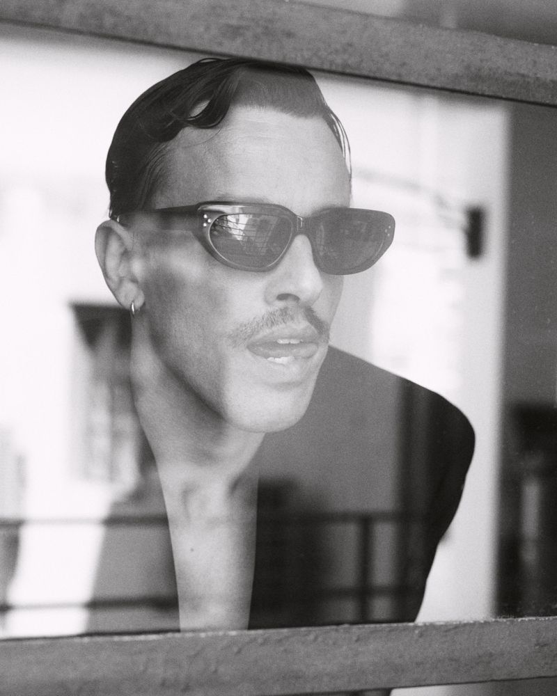 Portrait of a man wearing dark sunglasses and a mustache viewed through a reflective glass window for Spaghetti Magazine