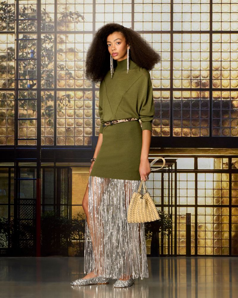 Model posing in an olive green knit dress with a metallic silver fringe skirt, holding a woven handbag in front of gridded glass.