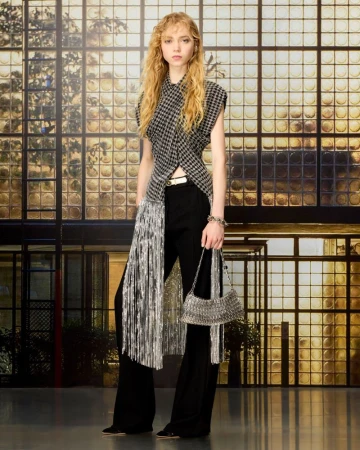 Model standing indoors wearing a patterned top with metallic fringe, black trousers, and holding a metallic handbag.