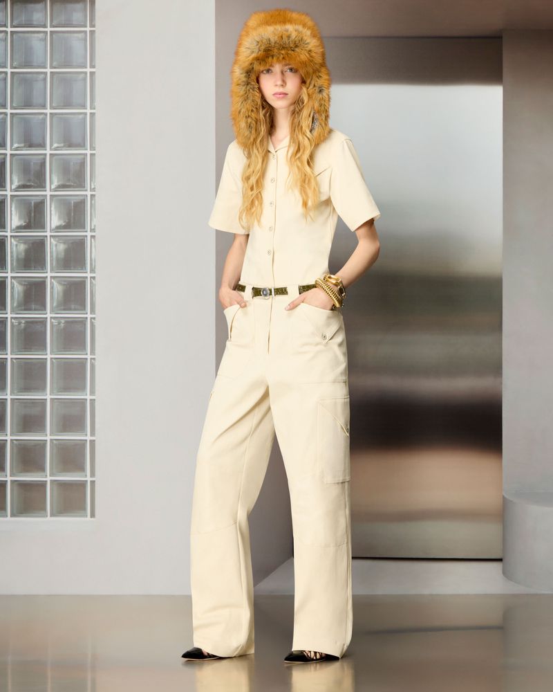 Model wearing an oversized brown fur hat and cream short-sleeved jumpsuit with hands in pockets in a minimalist studio setting.