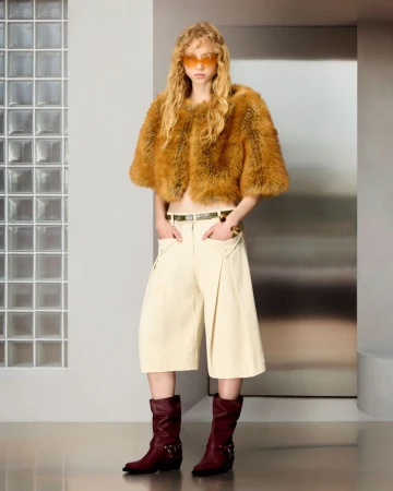Model in a cropped caramel fur jacket, wide cream culottes, and dark burgundy boots against a glass block studio wall.