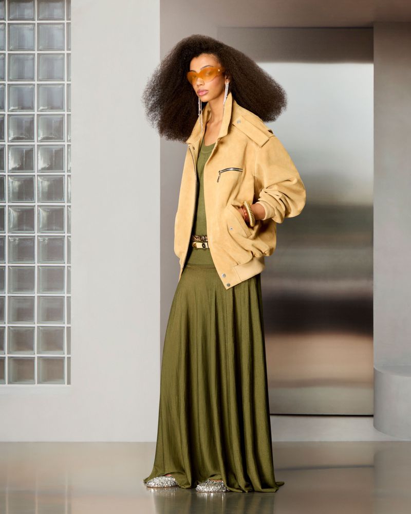 Model with voluminous afro hair in an oversized beige jacket and long olive green dress standing by a glass block wall.