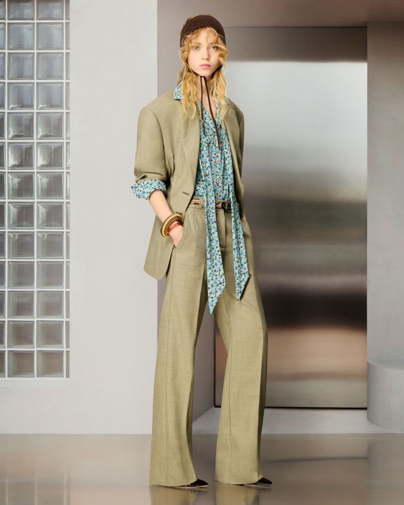 Model posing in an olive green tailored suit with wide-leg trousers, a blue floral neck-tie blouse, and a brown headpiece.