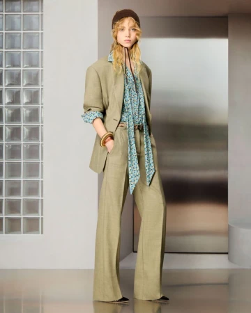Model posing in an olive green tailored suit with wide-leg trousers, a blue floral neck-tie blouse, and a brown headpiece.